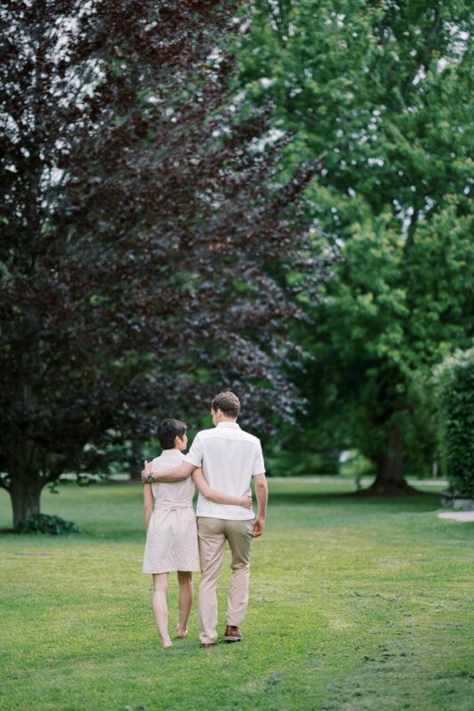 A couple walking in a lush, green park with tall trees, enjoying a serene afternoon together; the man has his arm around the woman, creating a warm, romantic atmosphere.
