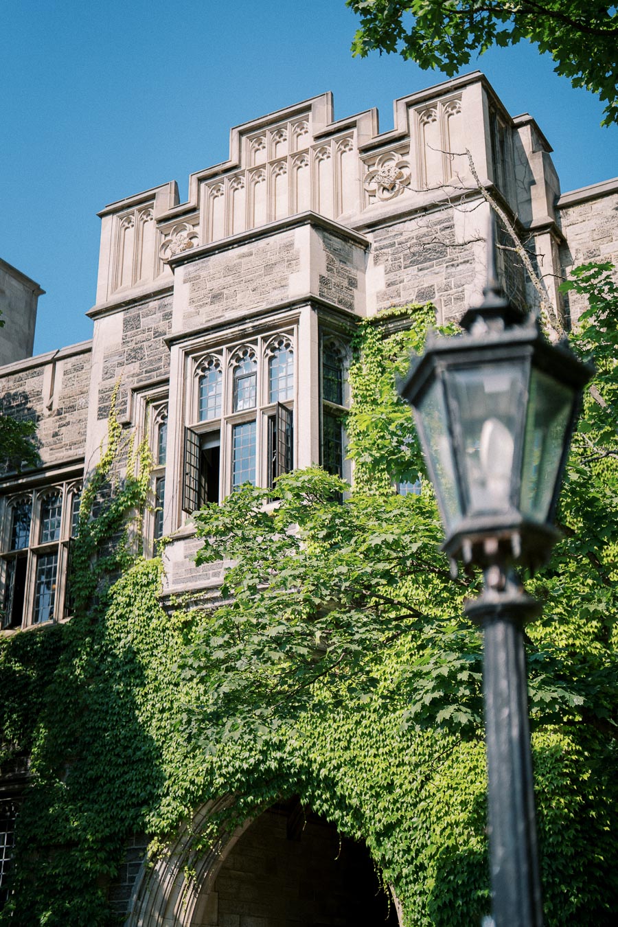 Historic stone building covered in green ivy with ornate arched windows and a vintage lamppost in the foreground under a clear blue sky.