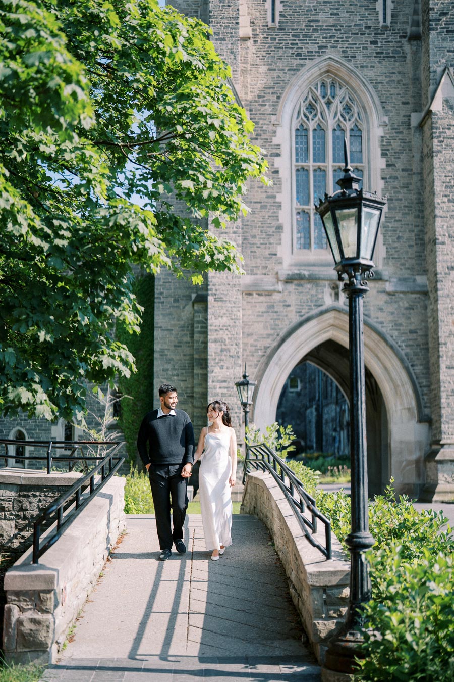 A couple walking hand in hand on a sunny day, surrounded by lush greenery, in front of a historic stone building with arched windows and vintage street lamps.