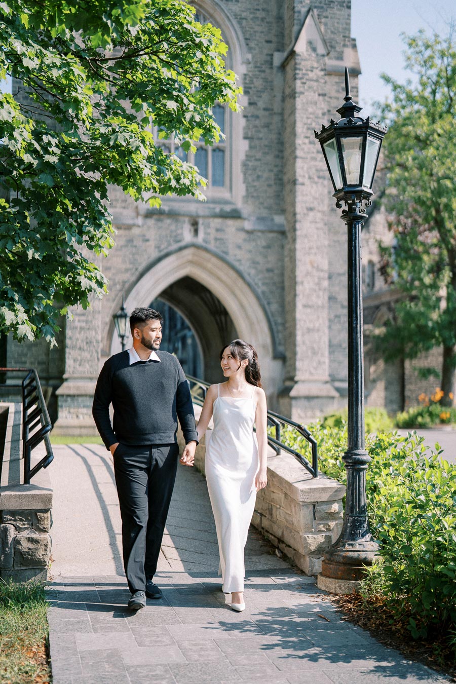 A couple holding hands and walking in front of a historic stone building, with lush greenery and an iron lamppost.