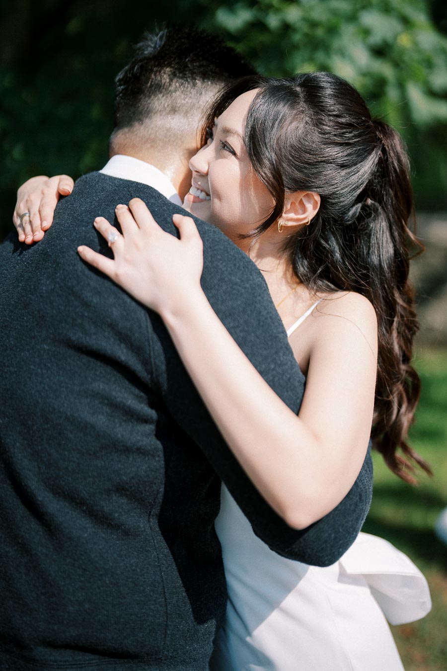 Young couple embracing outdoors on a sunny day, woman smiling in a white dress, green foliage in the background