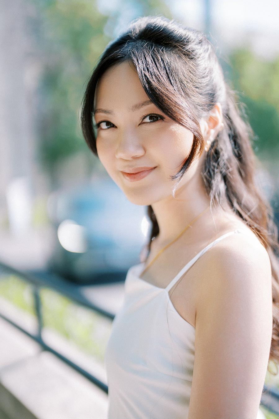 Young woman in a white dress standing outdoors, smiling softly with a blurred background of nature.