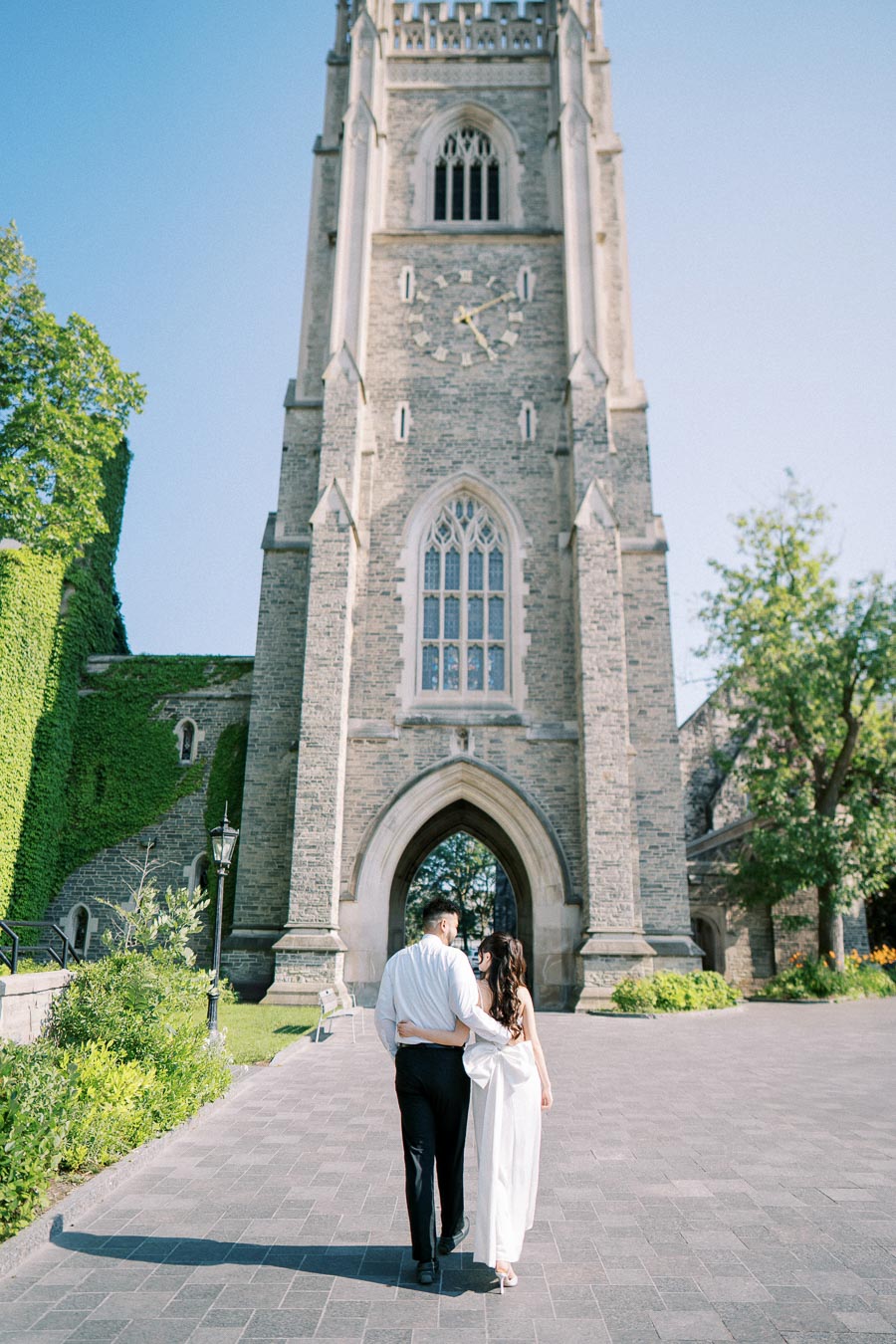 A couple walking arm-in-arm towards a historic stone tower on a bright, sunny day, surrounded by lush greenery.
