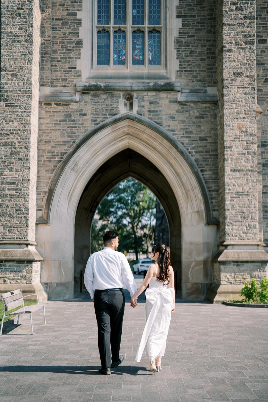 Couple holding hands walking towards a historic stone archway, wearing formal attire, with a backdrop of beautiful stained glass windows above.