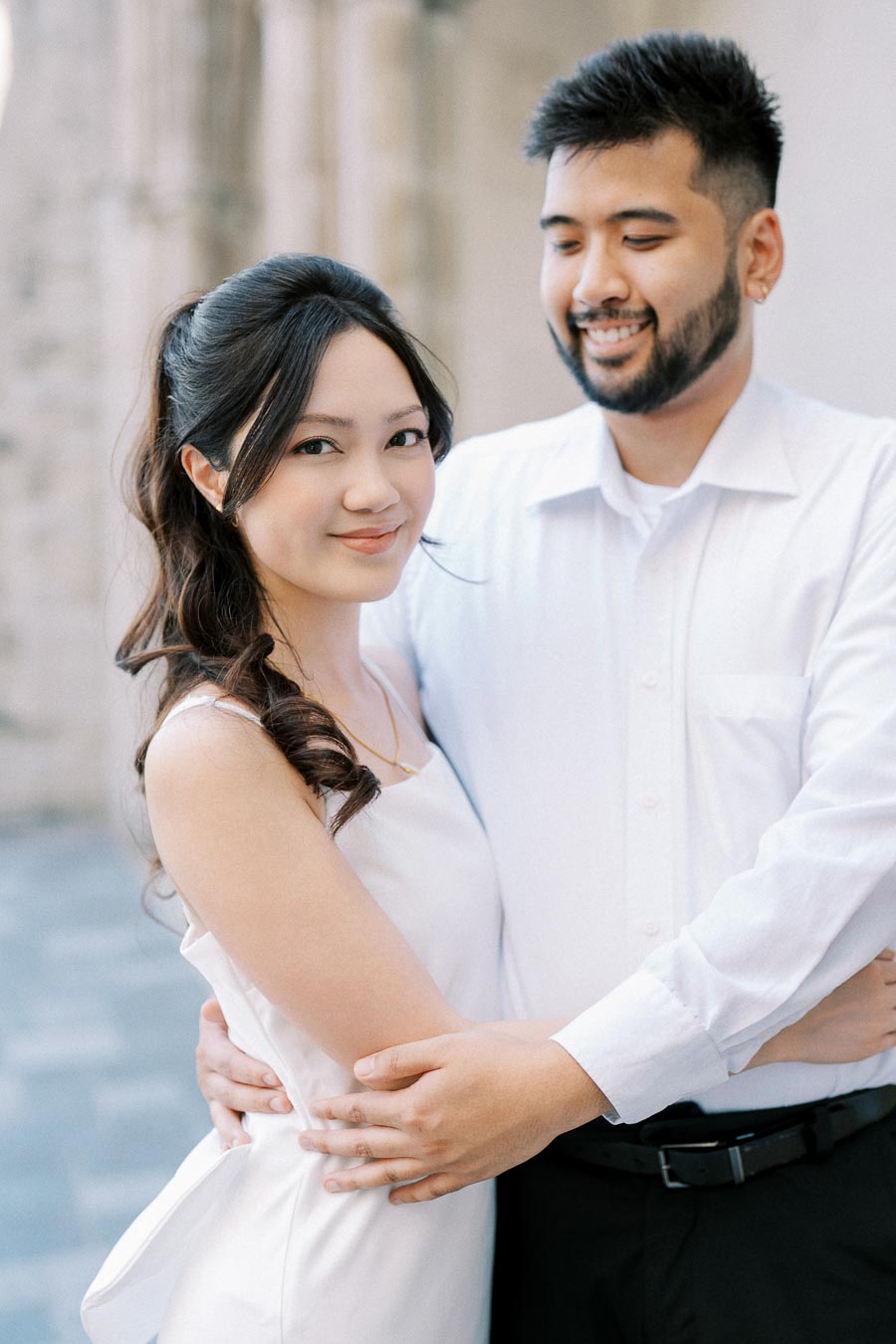 A couple embracing in a romantic outdoor setting, both smiling and wearing white attire, symbolizing love and happiness.