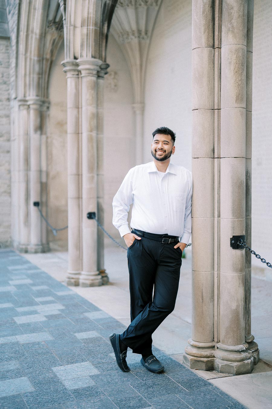 A man in formal attire leans against a stone column in a historic architectural setting, smiling confidently.