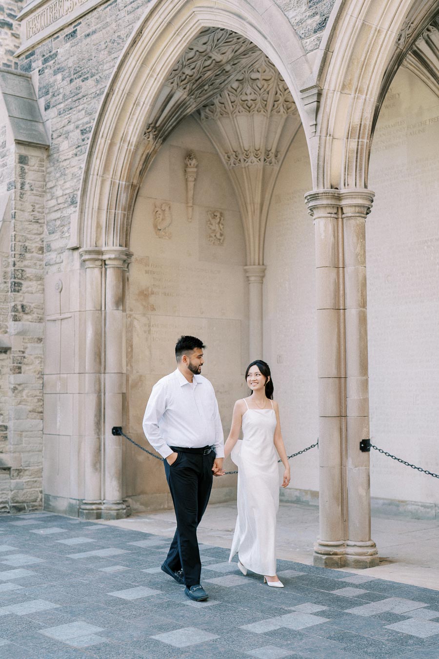 A couple holding hands and smiling while walking under a historic stone archway, dressed elegantly in formal attire.