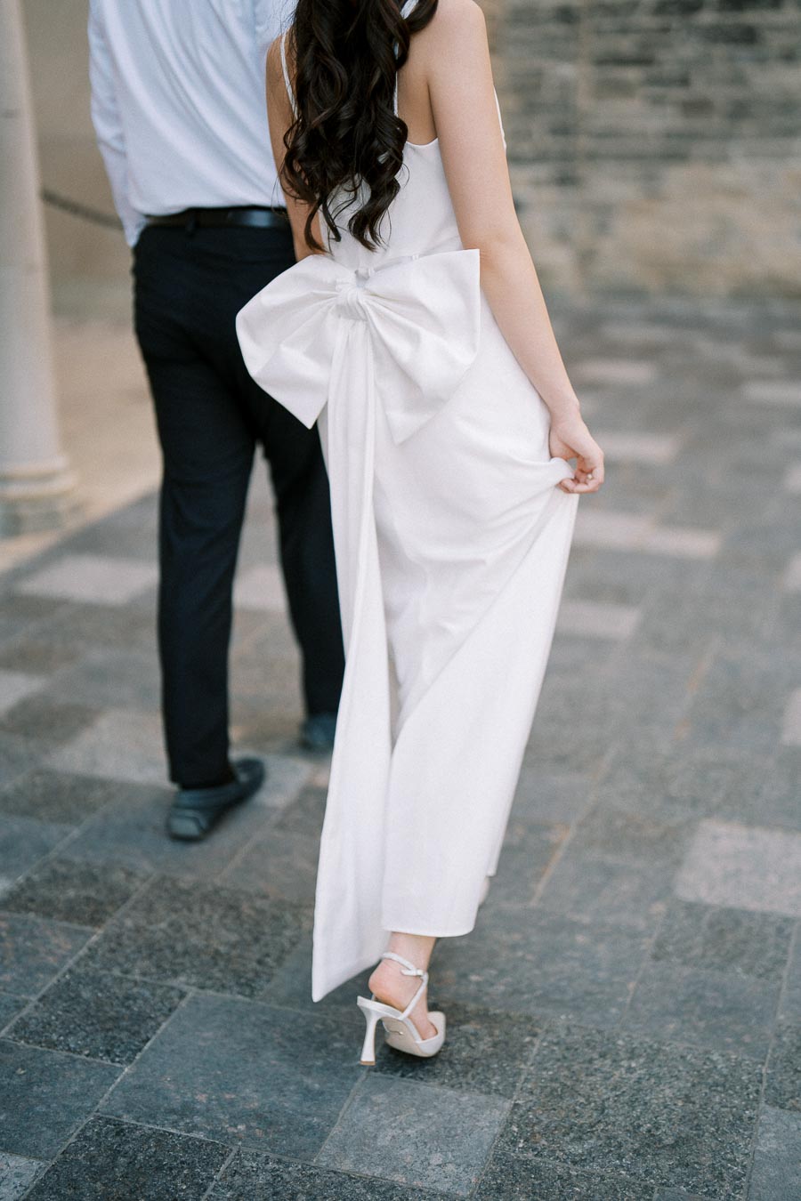 A woman in a white dress with a large bow at the back walks on a stone pathway, holding the hem of her dress, while accompanied by a man in formal attire.