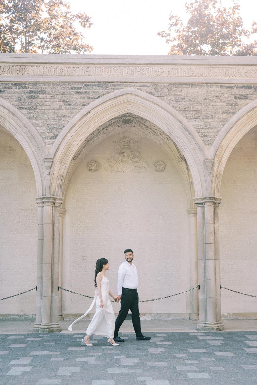 A couple holding hands and walking under historic stone arches with intricate carvings, surrounded by sunlight and trees.
