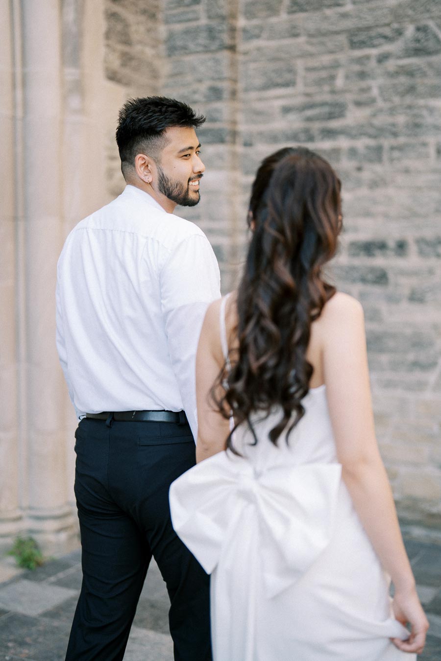 A couple dressed elegantly in semi-formal attire, walking together near a stone building. The man is wearing a white shirt and black pants, while the woman has long hair and a dress with a large bow at the back.