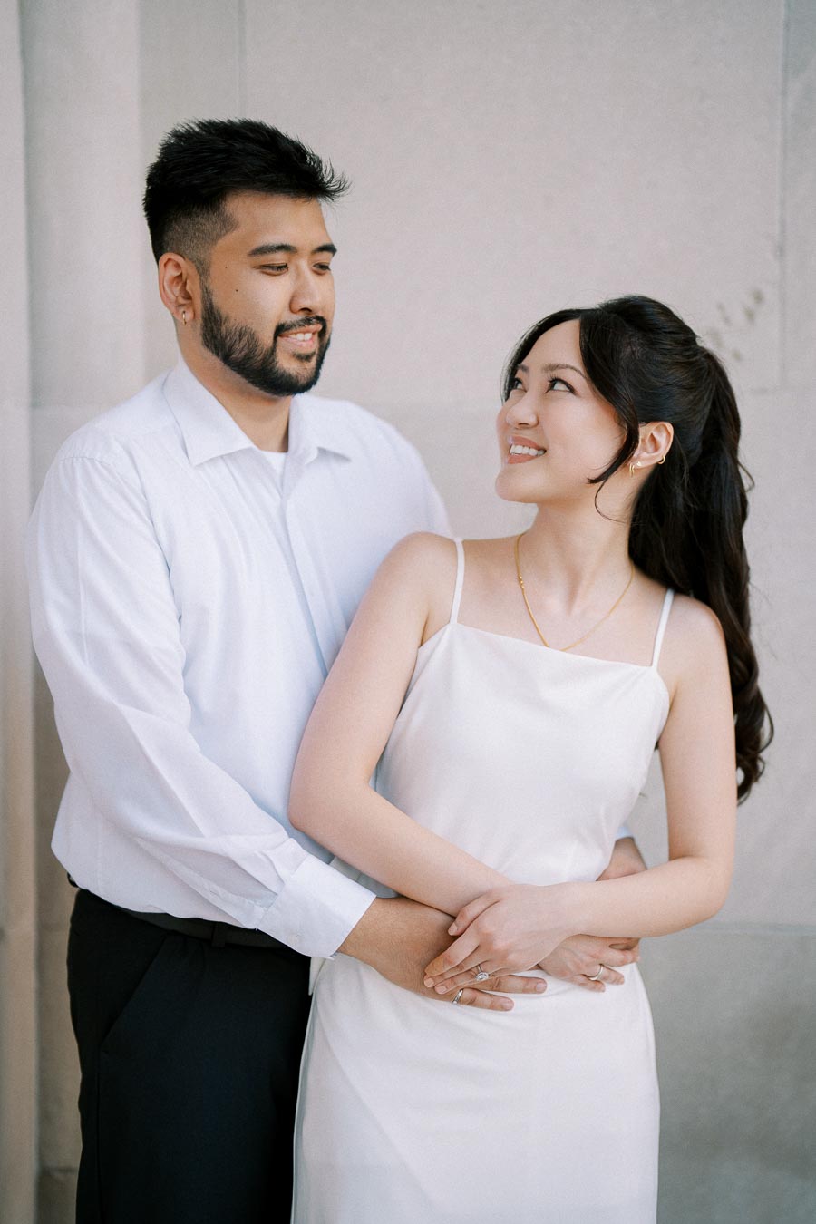 A couple smiling and embracing while standing against a light-colored wall, capturing a romantic moment.