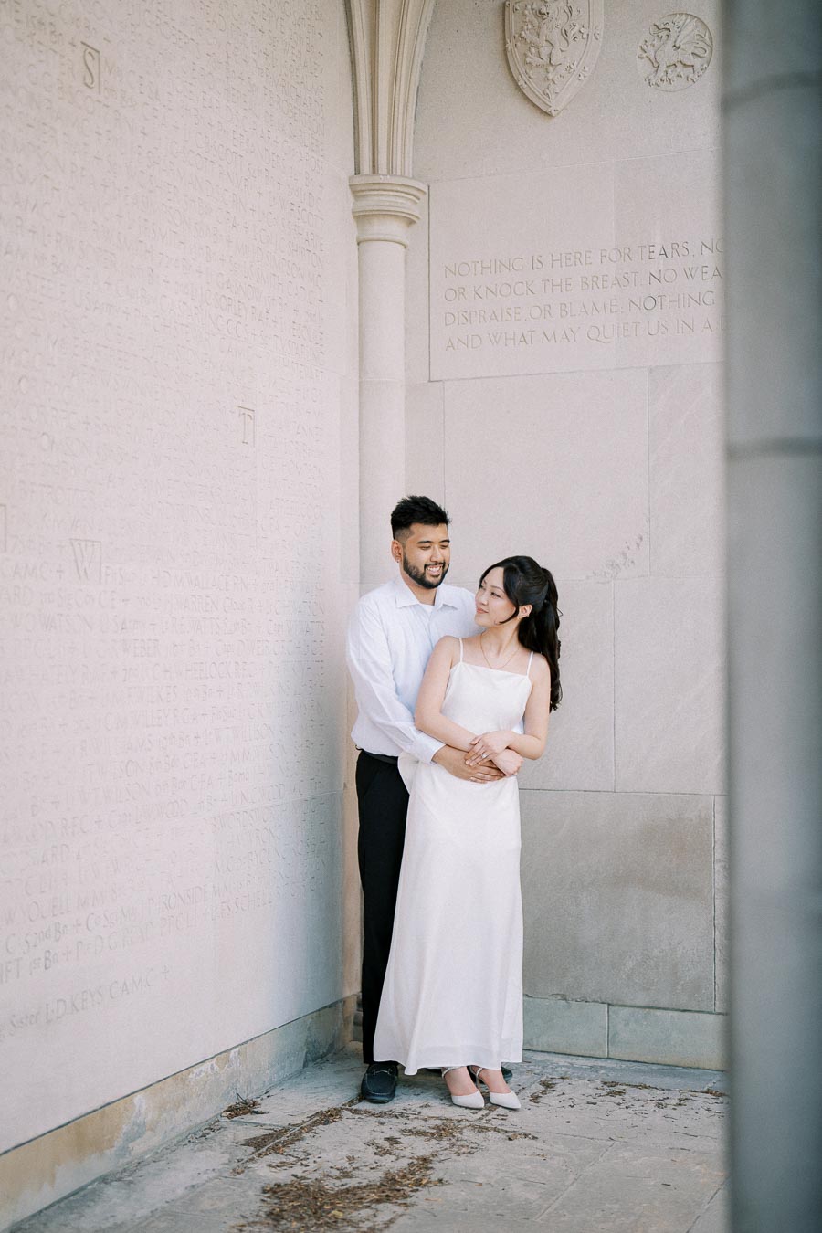 A loving couple embraces against a stone wall with engraved text in a serene setting, capturing a romantic moment.