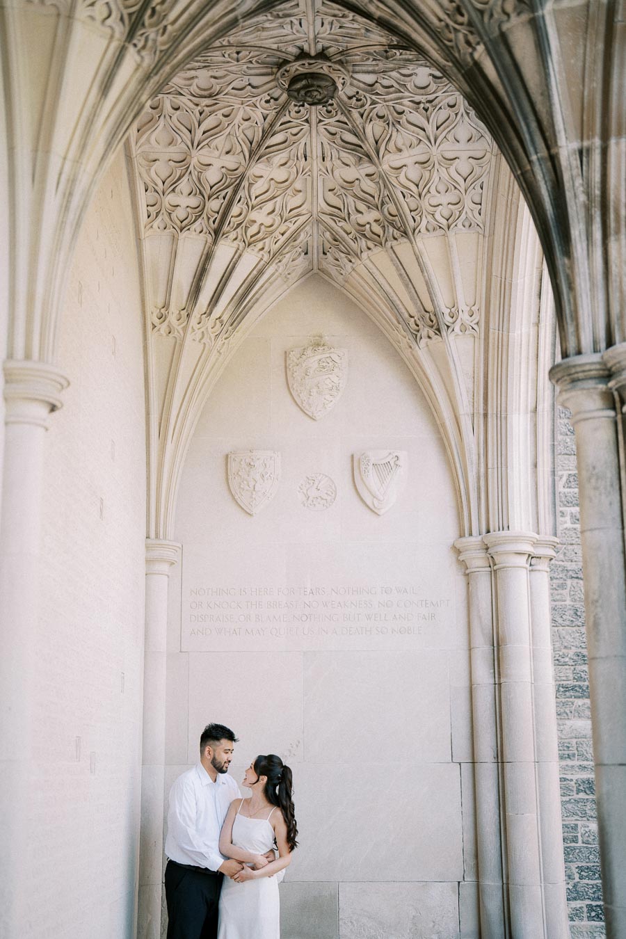 A couple embracing under ornate stone arches with intricate carvings and emblems on the wall behind them.