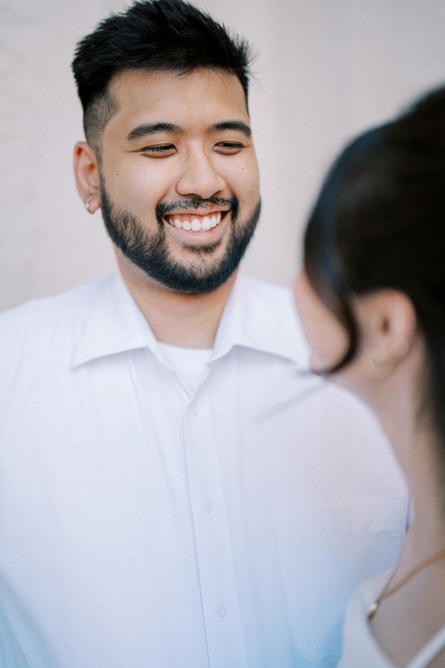 A man with a beard smiling warmly at a woman, wearing a white shirt and standing against a light background.