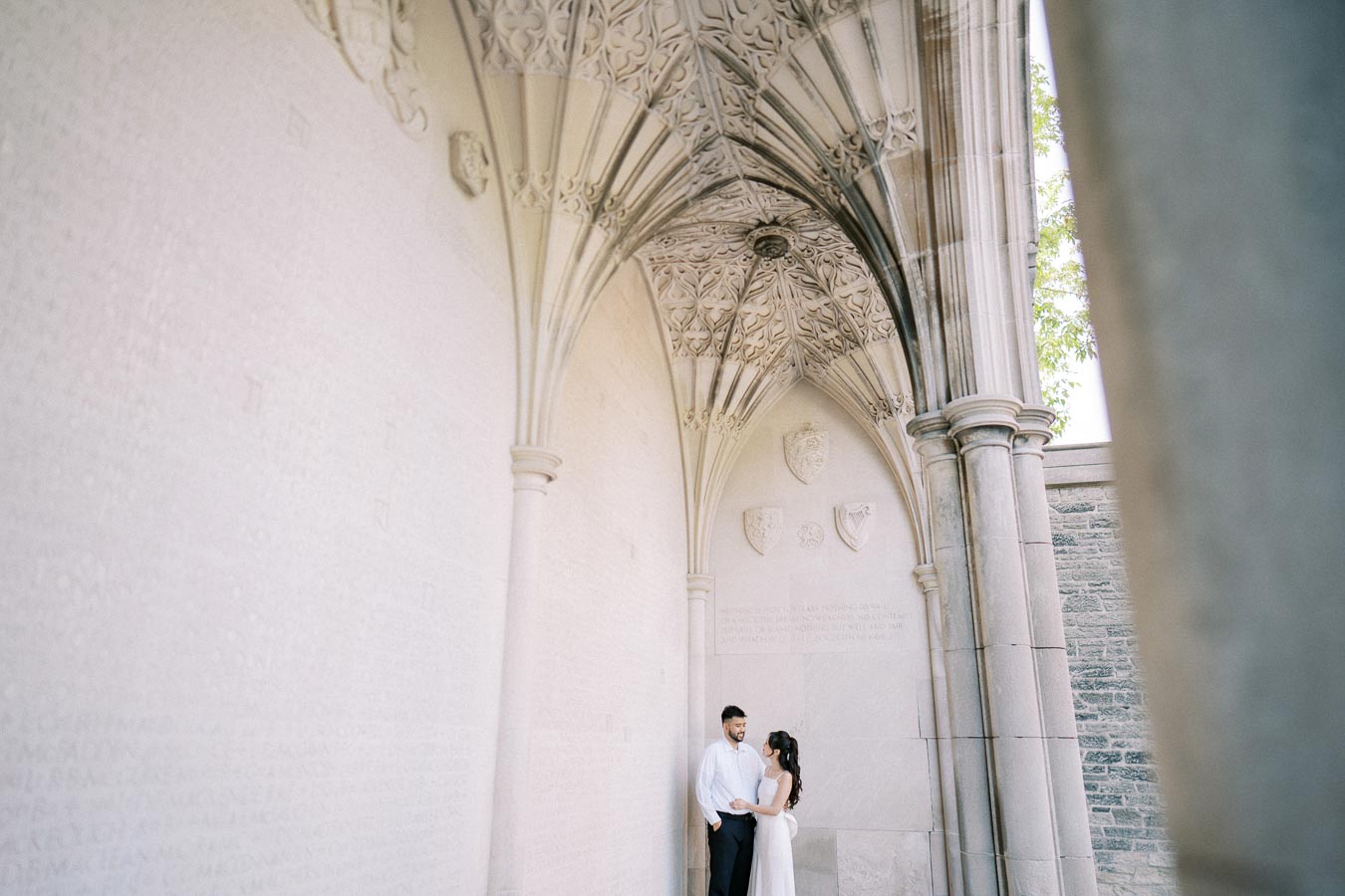 A couple in formal attire standing under a decorative arched stone ceiling with intricate carvings and emblems, in a historic architectural setting.