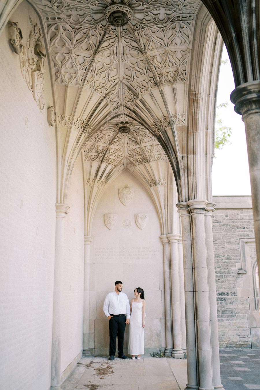 A couple stands beneath a decorative stone ceiling with elaborate Gothic architecture, intricately carved arches, and shield motifs, set against a serene and historic stone wall background.