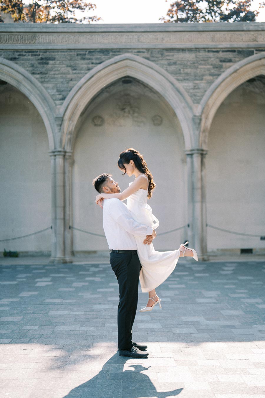 A couple in formal attire sharing a romantic moment outdoors, with the man lifting the woman in front of an elegant stone archway.