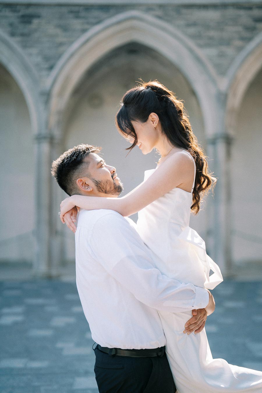 A couple in formal attire embraces under an arched stone building, capturing a joyful romantic moment.