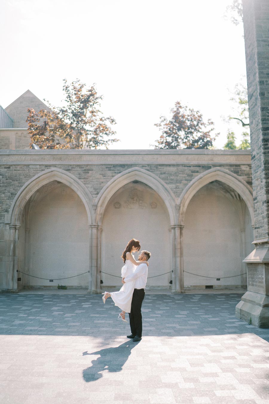 A couple embracing in a romantic courtyard setting, with arched stone architecture and sunlight filtering through, creating a warm and timeless atmosphere.
