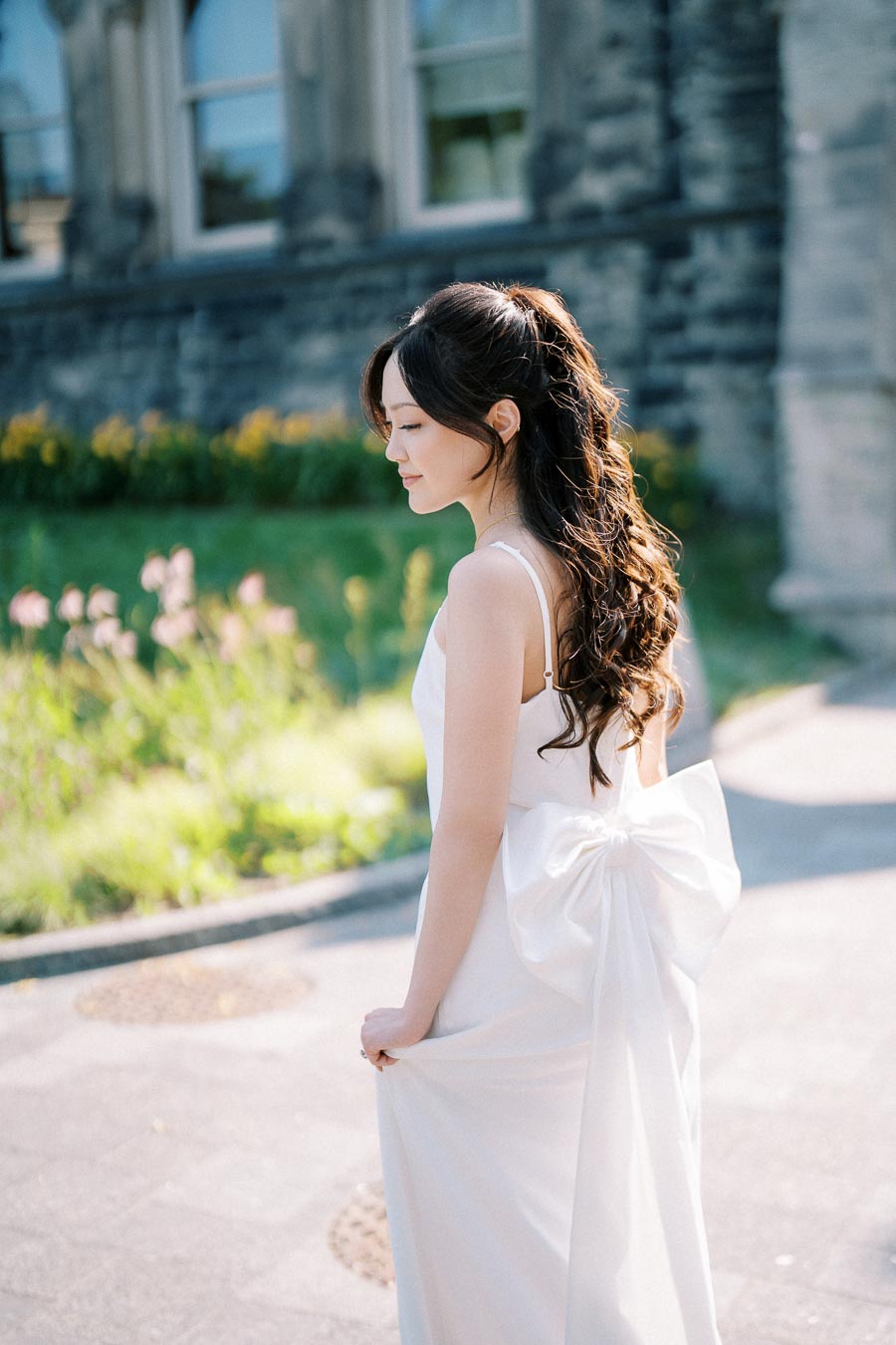 A bride in a white wedding dress with a large bow on the back stands outdoors, surrounded by greenery and a stone building, capturing a serene and elegant moment.