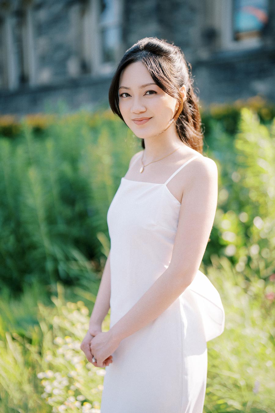 A woman in a white dress stands in a green garden setting, looking at the camera with a gentle smile, framed by blurred, lush foliage.