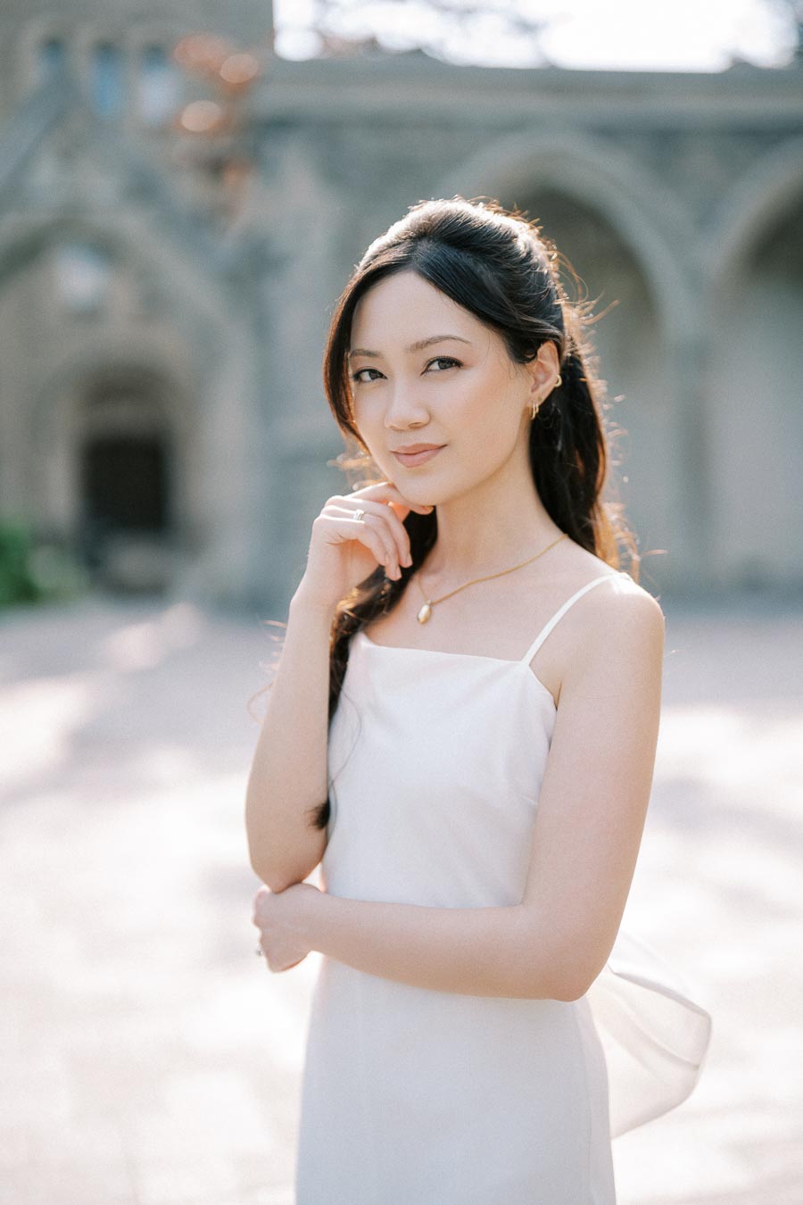 Portrait of a woman in a white dress standing outdoors, softly lit with a blurred architectural background, conveying elegance and serenity.