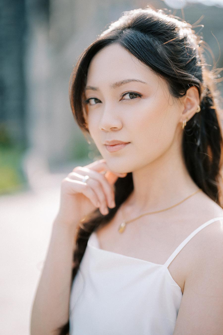 A woman with long dark hair wearing a white top and necklace, posing outdoors with a thoughtful expression, surrounded by soft natural light.