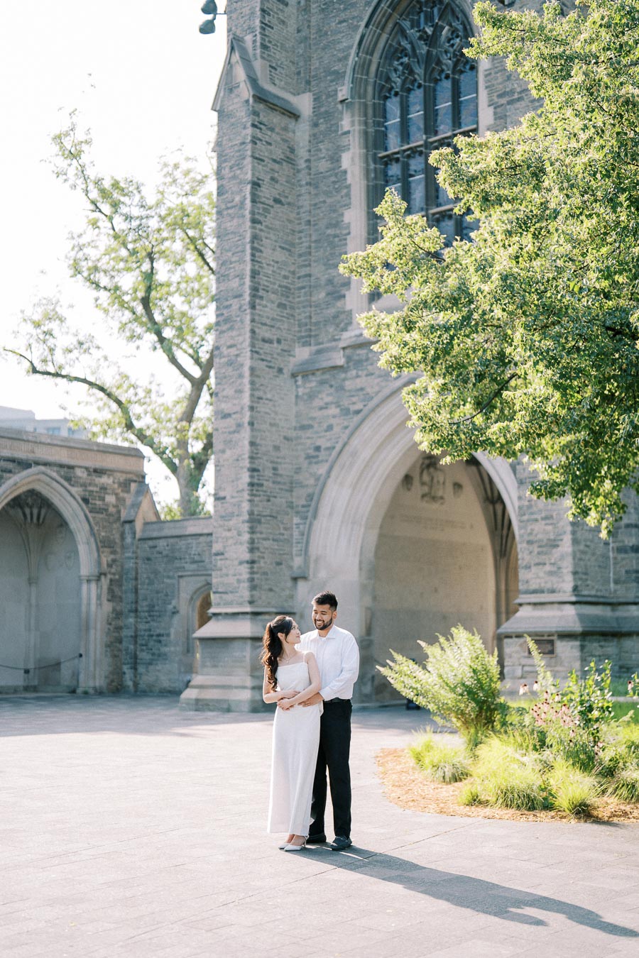 A happy couple embracing in front of a historic stone building with arched windows, surrounded by greenery on a sunny day.