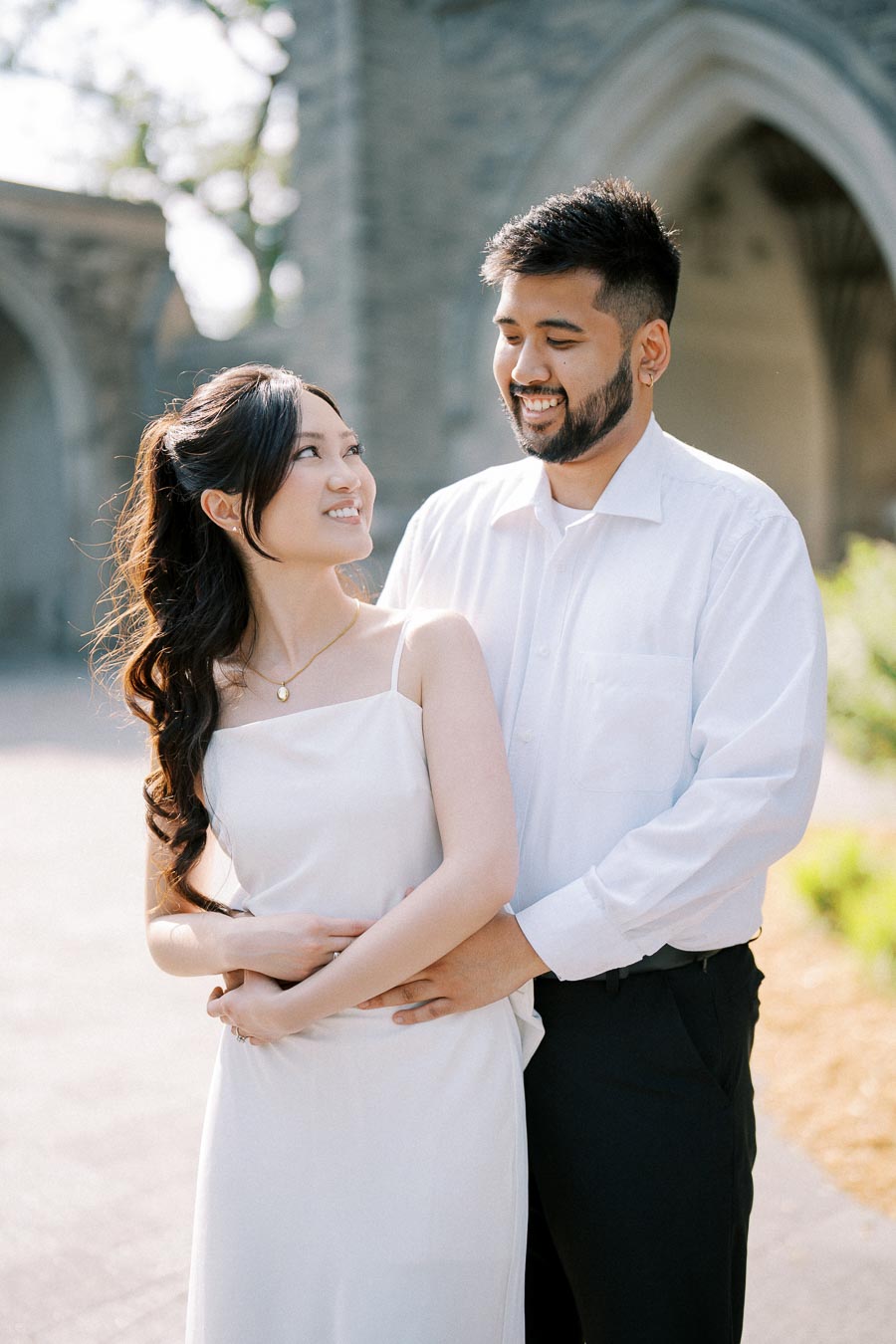 A happy couple embracing and smiling at each other in front of a historic stone building, both dressed in white outfits, with sunlight creating a warm, joyful atmosphere.