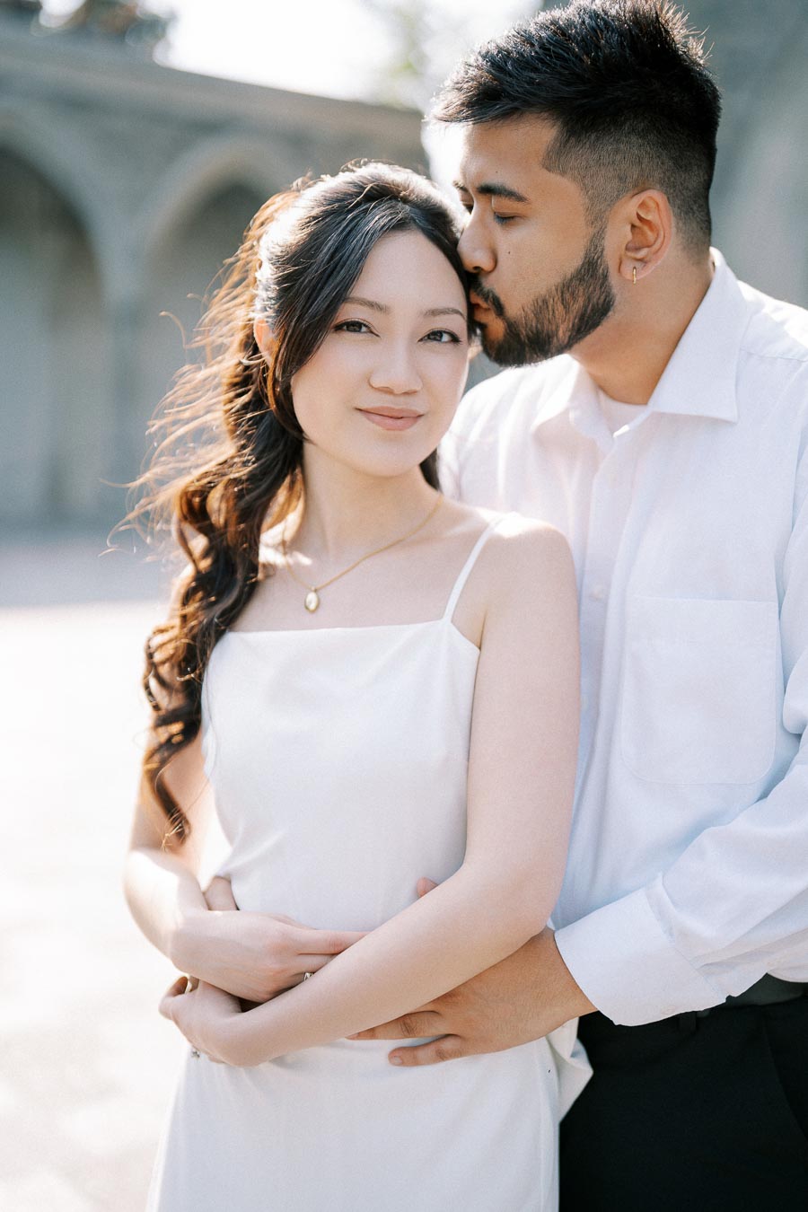 Romantic couple embracing outdoors, with the man gently kissing the woman's forehead, both dressed in elegant white attire, capturing a tender moment in natural light.