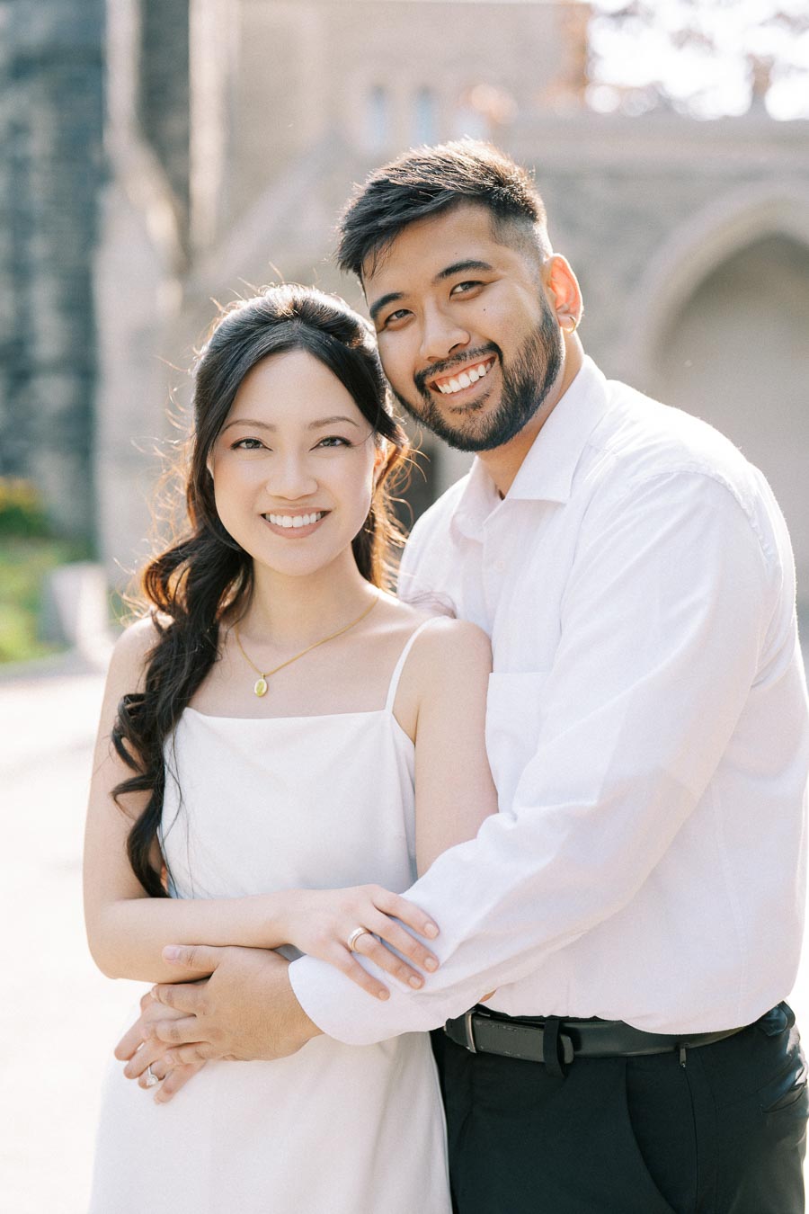 Smiling couple in white outfits embracing outdoors with an elegant background, symbolizing happiness and togetherness.