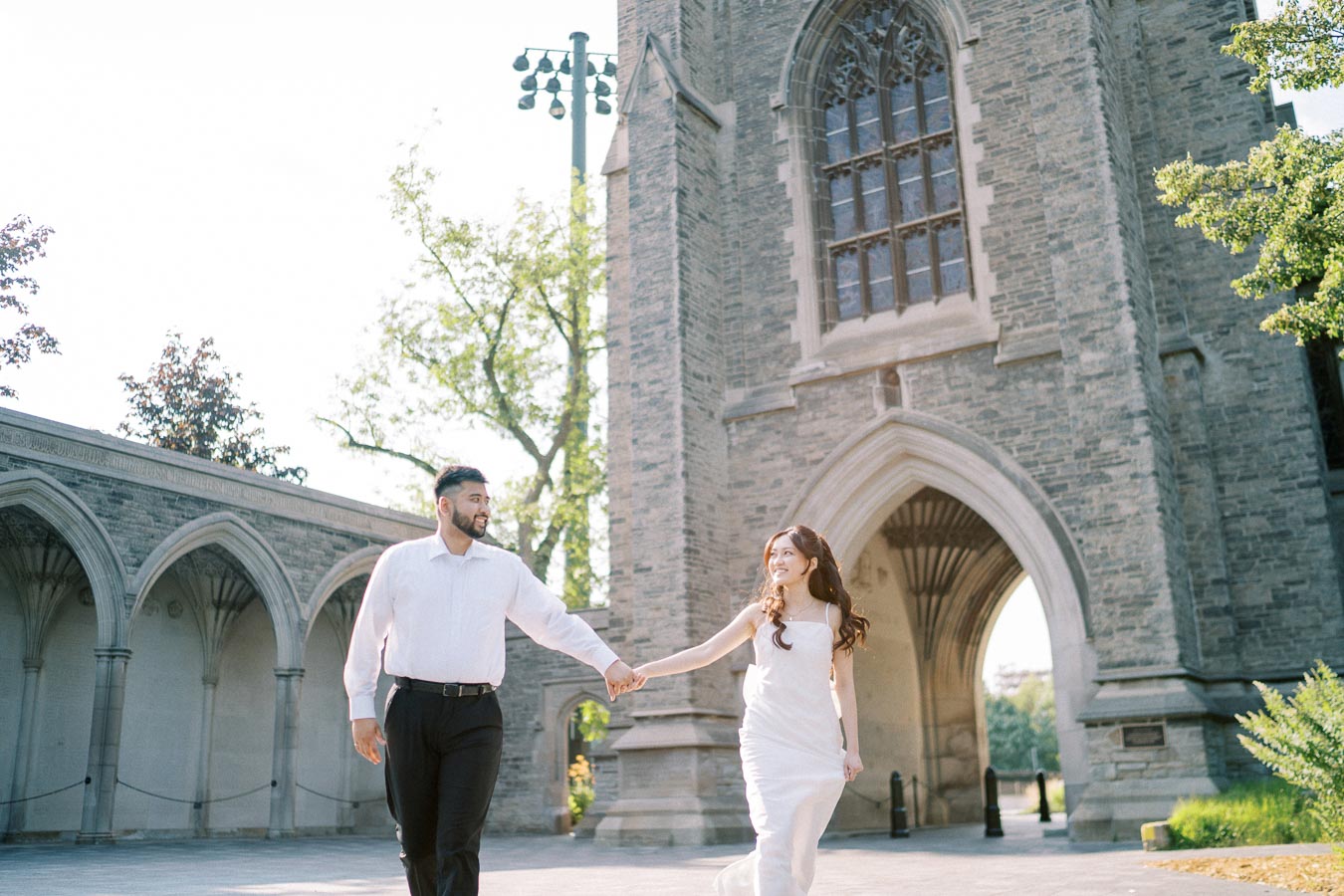 A couple walking hand in hand in front of a historic stone building with arched windows, surrounded by lush greenery on a sunny day.