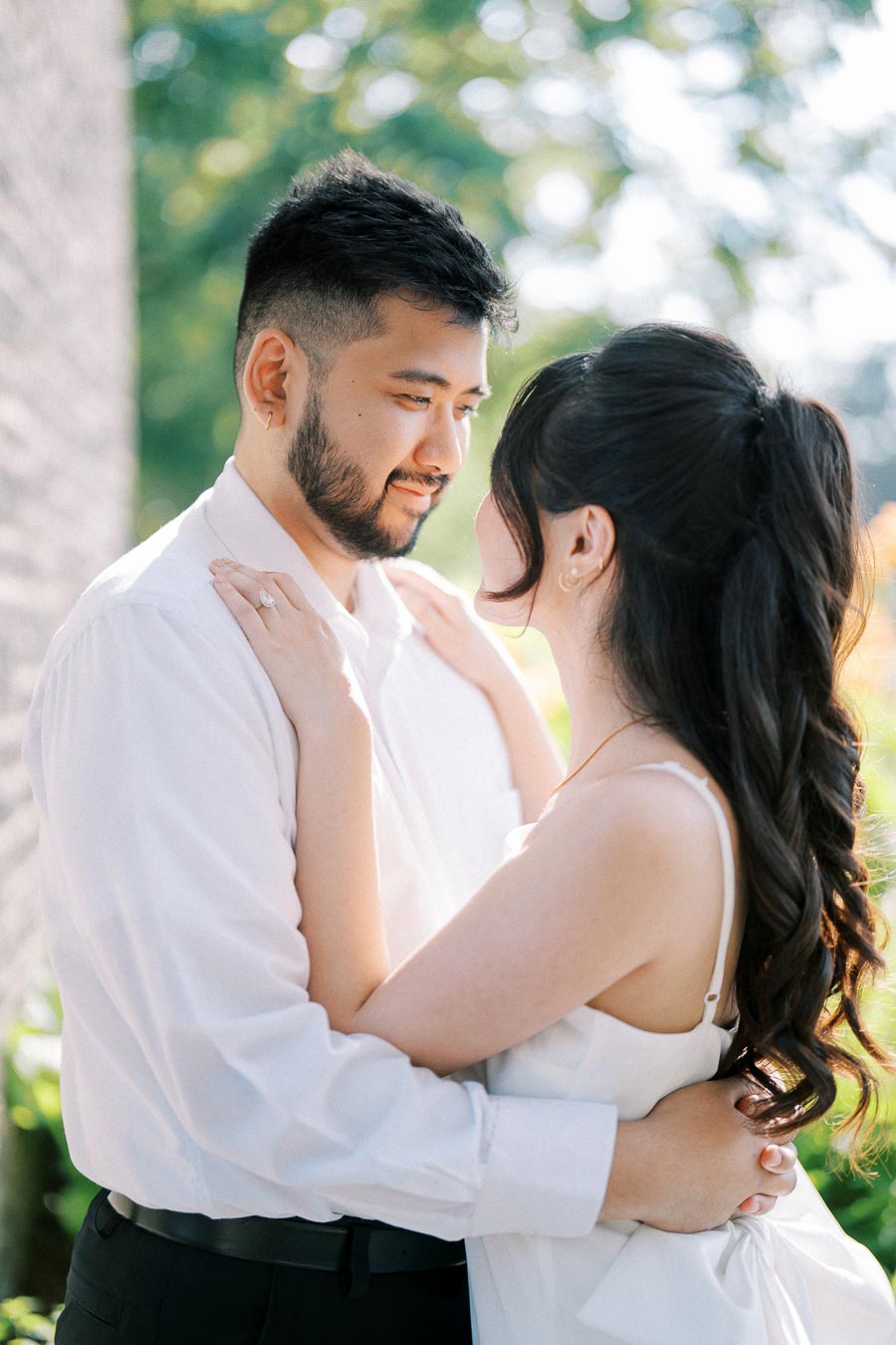 A couple in love, embracing outdoors surrounded by lush greenery and sunlight, wearing white attire and appearing joyful and affectionate.