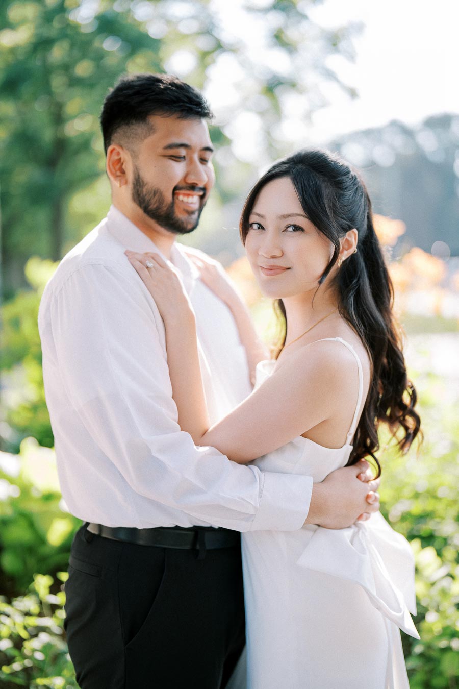 Romantic couple embracing in a sunlit garden setting, both dressed in formal white attire and sharing a joyful moment.