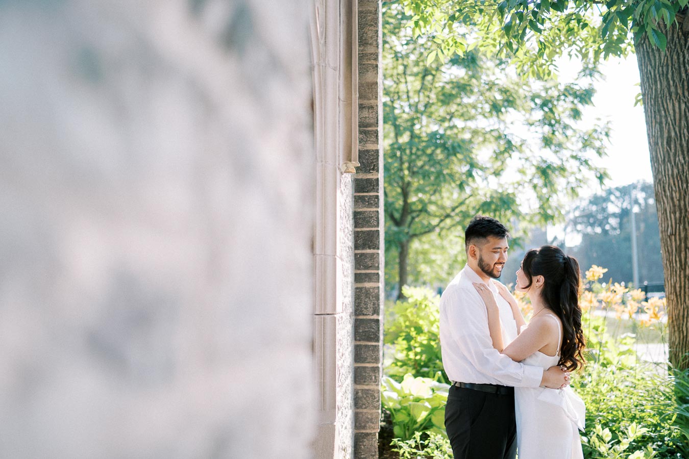 A couple embraces lovingly in an outdoor setting, surrounded by lush greenery and sunlight filtering through the trees, creating a romantic and serene atmosphere.