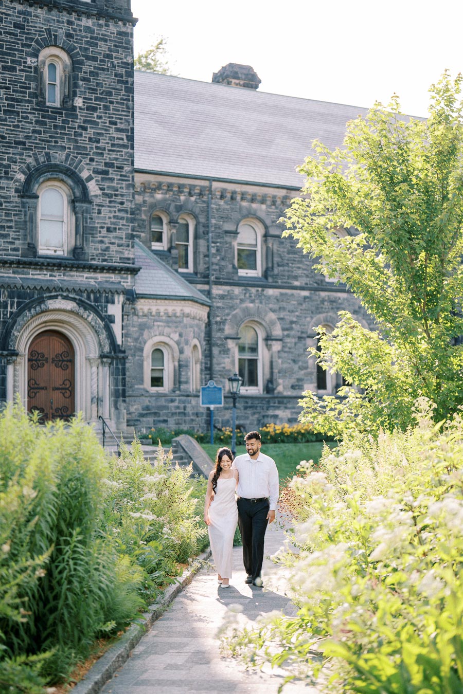 Couple walking on a garden path beside a historic stone building, surrounded by lush greenery and sunlight.