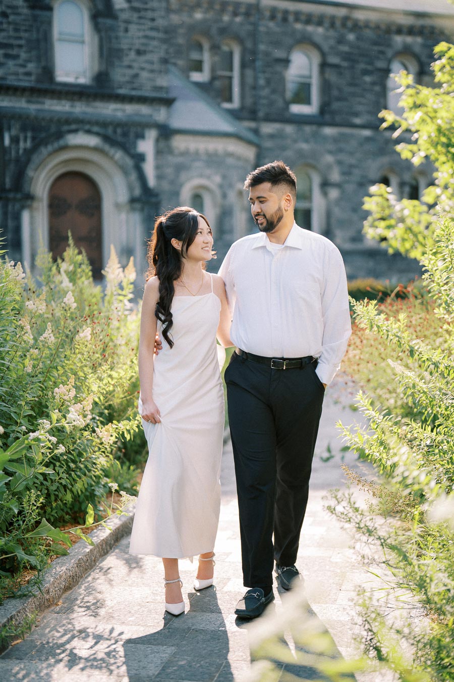 A couple walking arm in arm through a garden path, dressed in elegant white and black attire, with a historic stone building in the background, surrounded by lush green foliage on a sunny day.