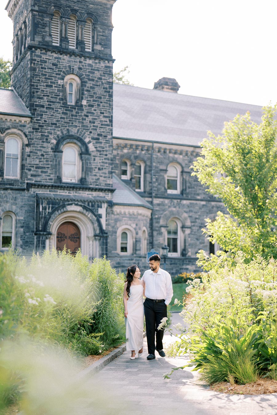 A couple walks arm in arm through a lush garden in front of a historic stone building, enjoying a sunny day.