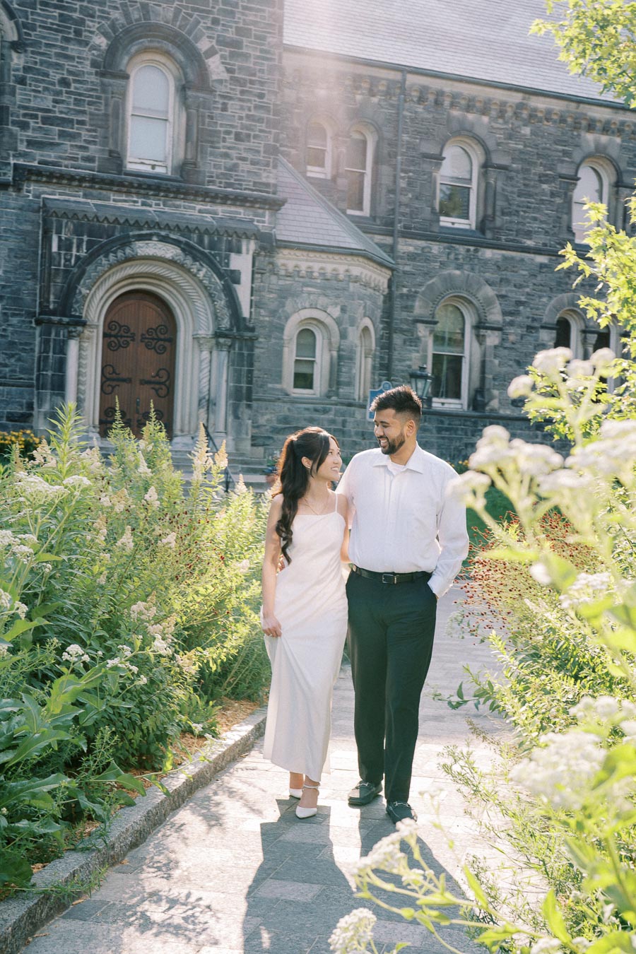 A couple walking arm in arm on a sunny path in front of a historic stone building, surrounded by lush greenery and flowers.