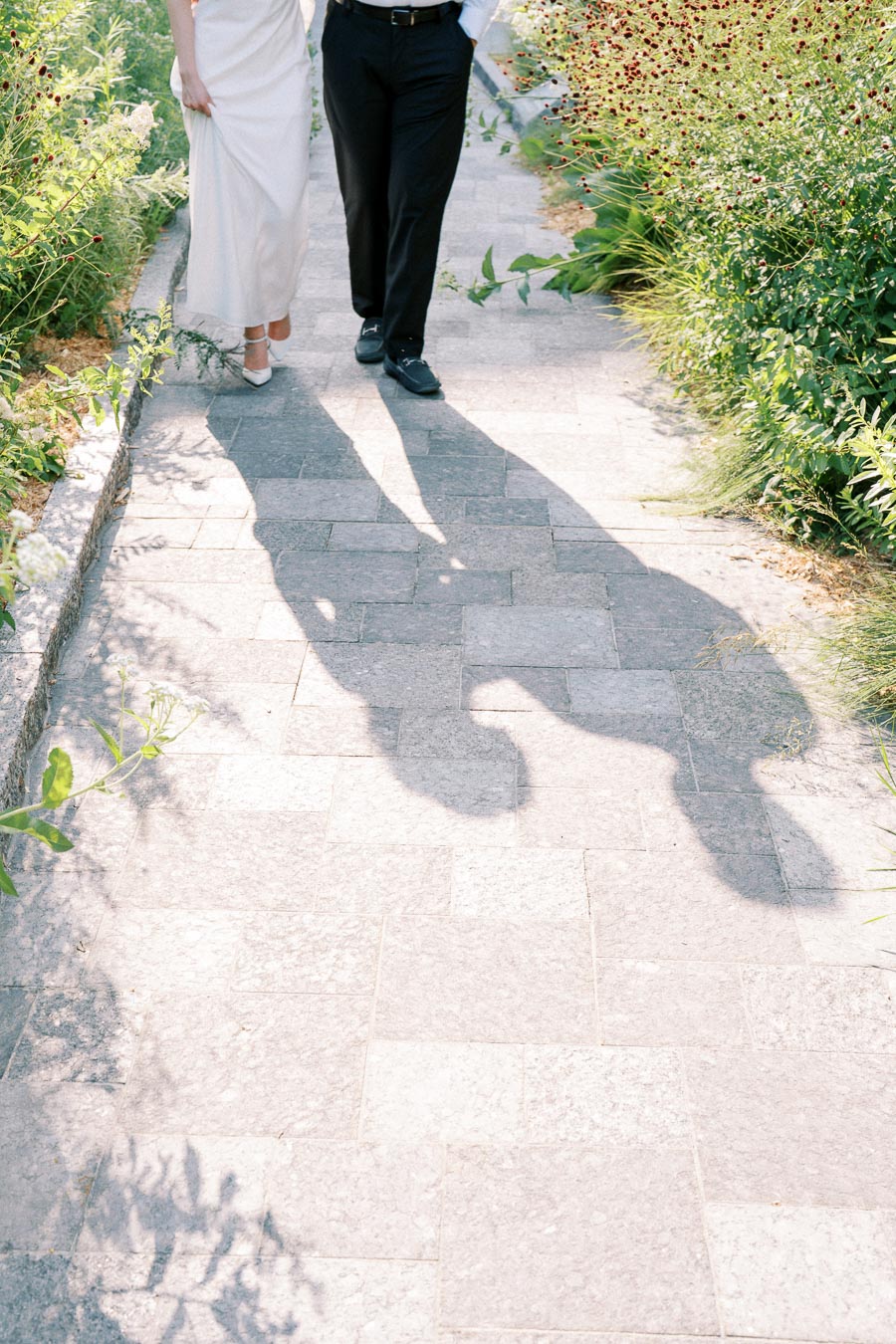 A couple walking hand in hand on a sunlit stone pathway, surrounded by lush greenery, casting long shadows on the ground.