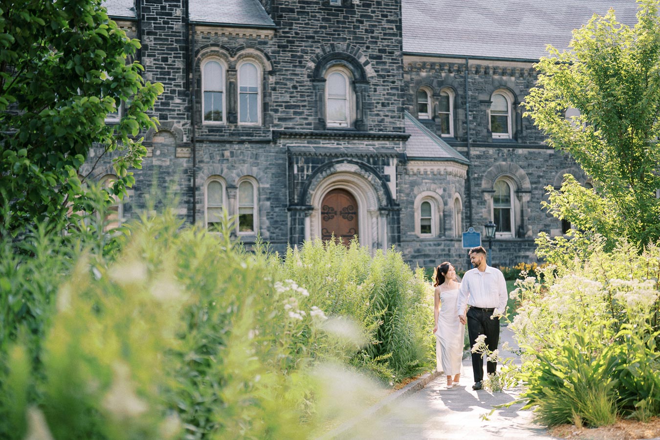 A couple holding hands and walking along a garden pathway in front of a historic stone building, surrounded by lush greenery and sunlight.