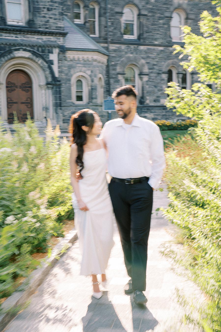 A couple walking arm in arm along a garden path, in front of a historic building.