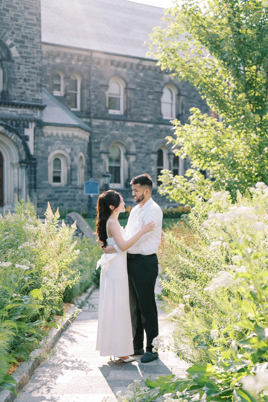 A couple embracing in a garden pathway with lush greenery and a historic stone building in the background, bathed in soft sunlight.