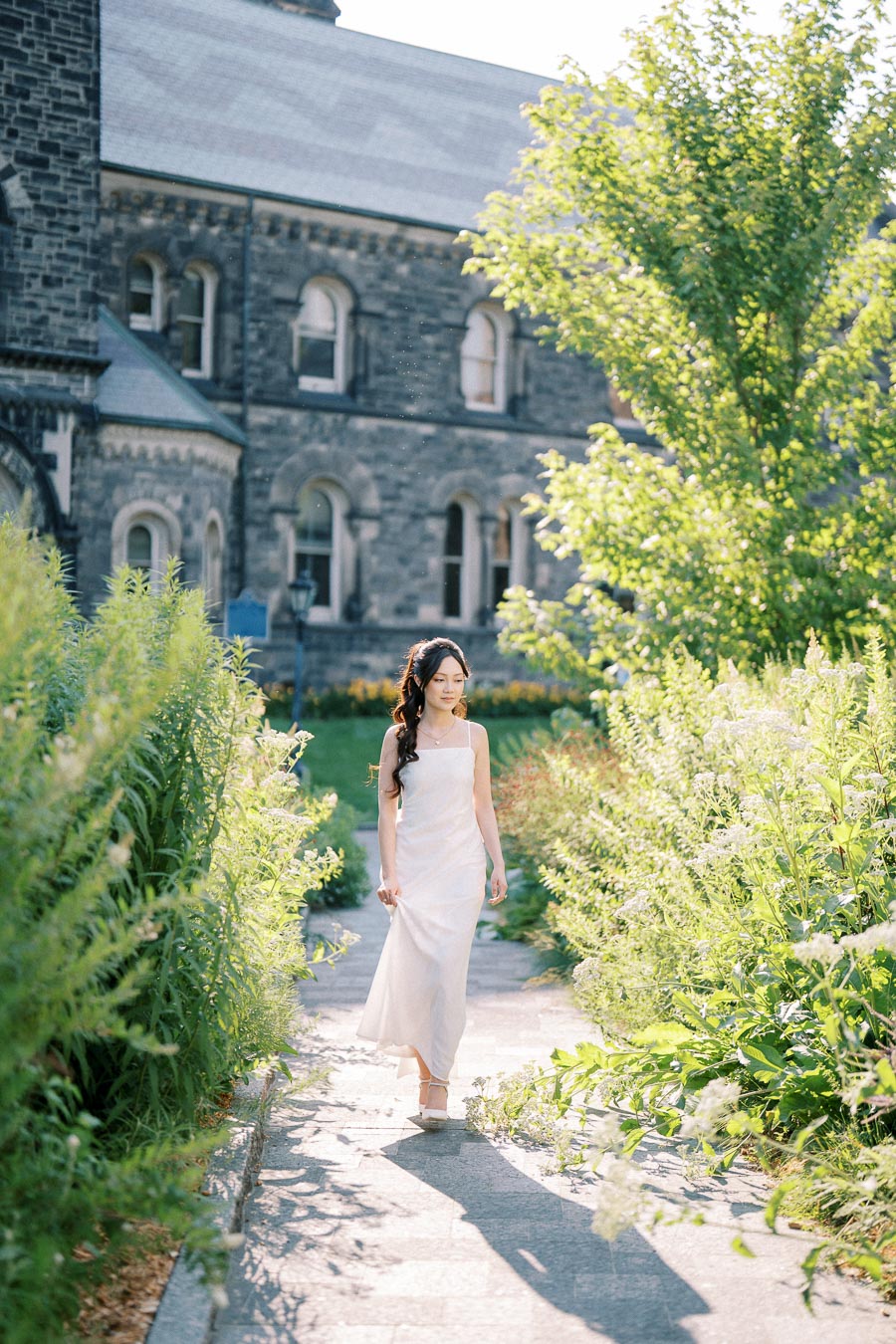 A woman in a white dress walks gracefully along a sunlit path, surrounded by lush green foliage, with a historic stone building in the background.