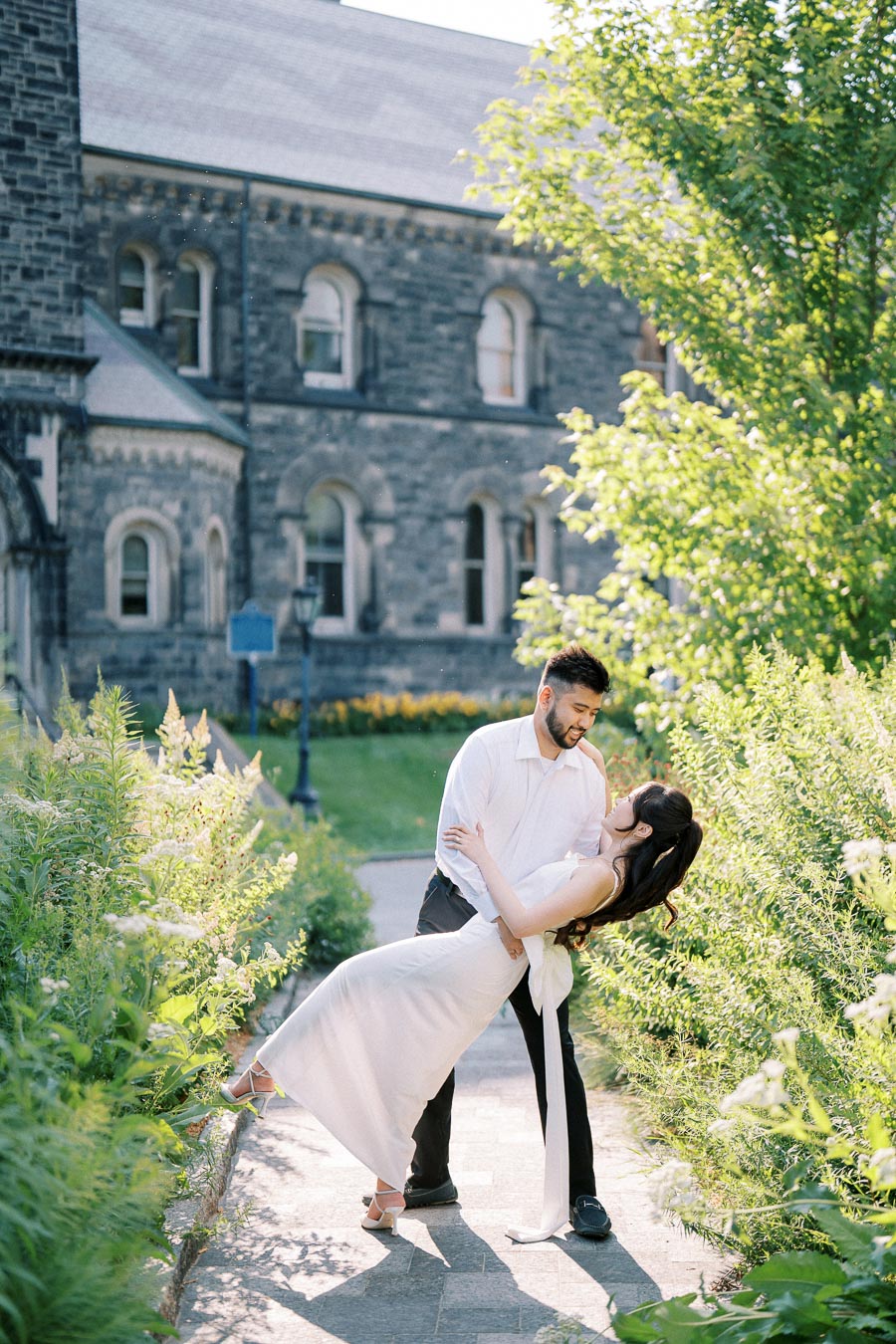 A couple in elegant attire shares a romantic dance in a lush garden setting, with a historic stone building in the background.