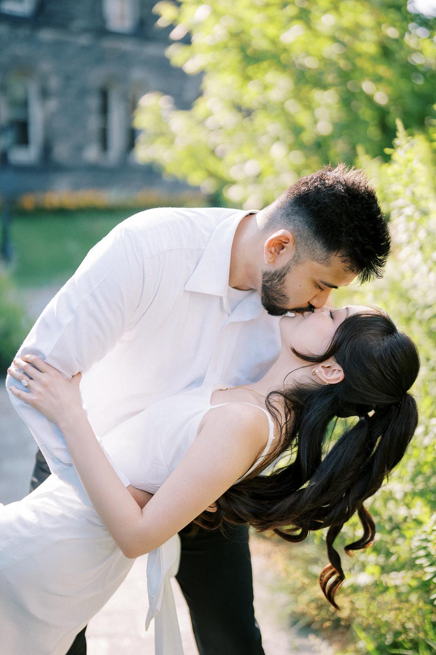 A couple kissing romantically in a garden setting, with lush greenery and a stone building in the background, both wearing white attire.