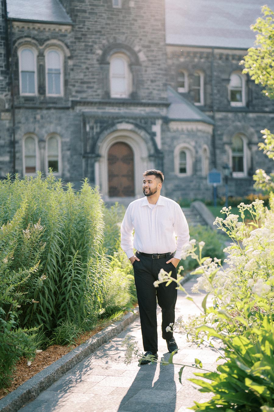 A person in a white shirt and black pants walks through a garden path with lush green plants, in front of a historic stone building with arched windows, on a sunny day.