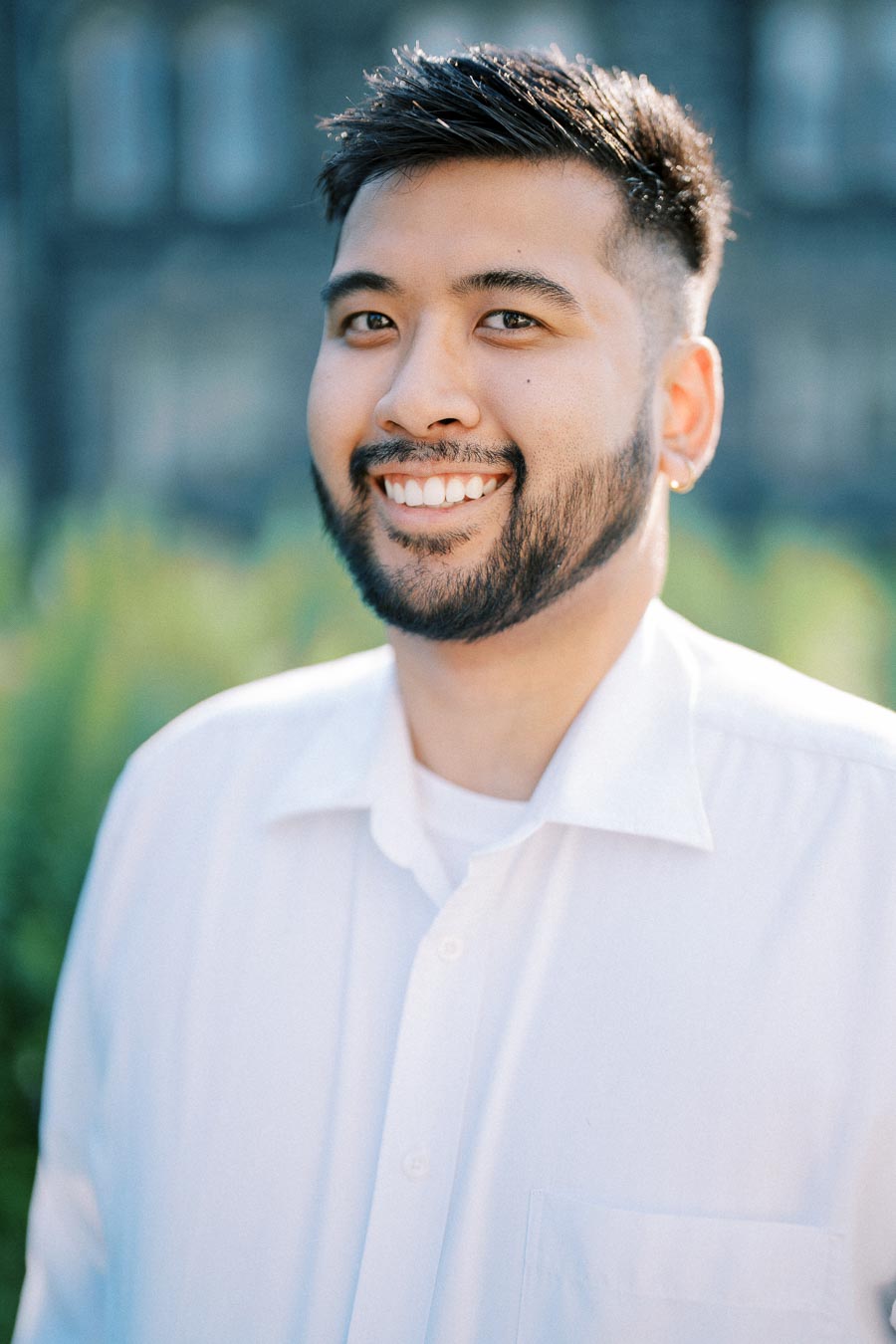 Portrait of a person smiling outdoors, wearing a white shirt, with greenery in the background.