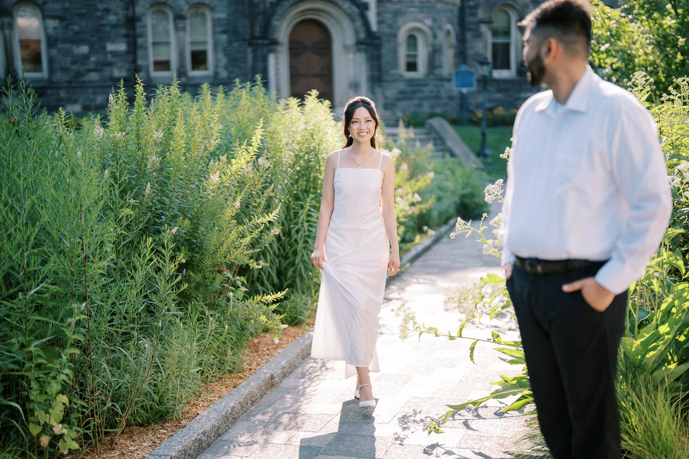 A woman in a white dress walking toward a man in a white shirt, surrounded by lush greenery and an old stone building in the background.