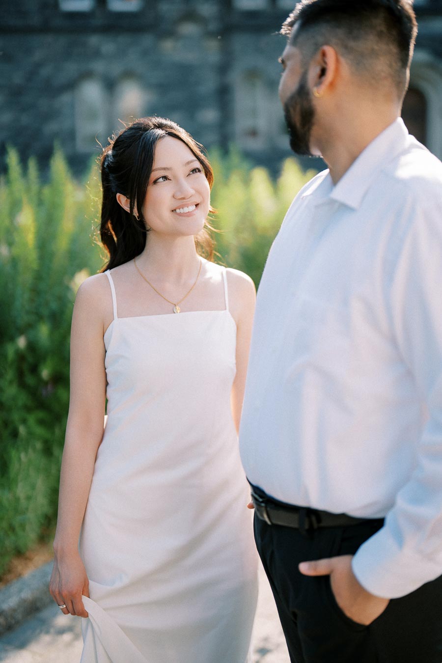 A couple dressed elegantly in white outfits, standing outdoors with a backdrop of greenery and a stone building, smiling at each other in a romantic setting.